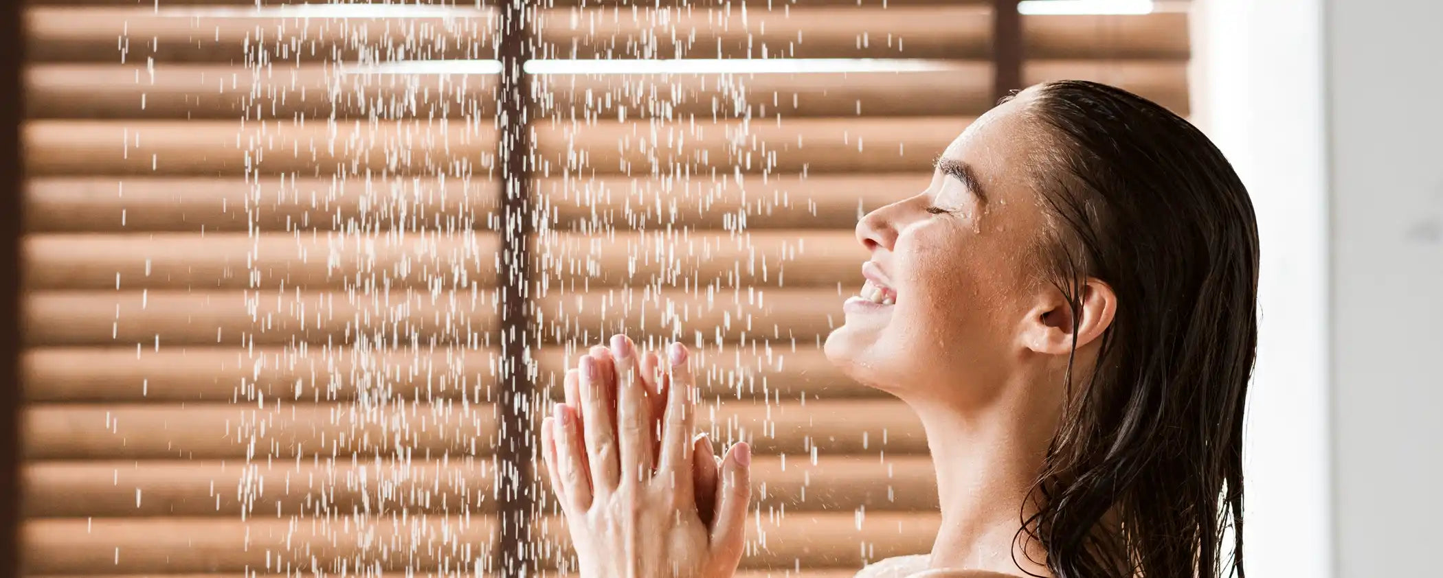 Woman standing under a Natural Action shower with water droplets falling, with wooden blinds in the background.
